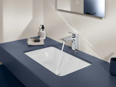 Un lavabo moderne avec une robinetterie Architectura Square Villeroy & Boch Un lavabo moderne avec de l’eau qui s’écoule, un distributeur de savon, un gant de toilette avec un morceau de savon et un miroir au-dessus. La tablette est bleue et un porte-brosse à dents se trouve à droite du lavabo.