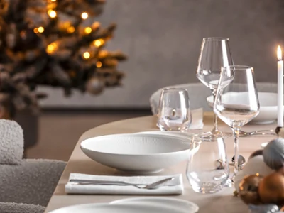 Afina, idéal pour la période de Noël Une table à manger dressée avec des verres et des assiettes Afina, à côté une bougie allumée et un sapin de Noël décoré en arrière-plan.