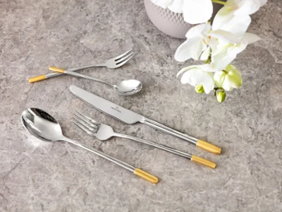 Un ensemble de couverts de table Ella de Villeroy & Boch avec des notes dorées Un élégant ensemble de couverts de table Ella avec des touches dorées sur un plateau de table en marbre, accompagné d’une orchidée blanche.
