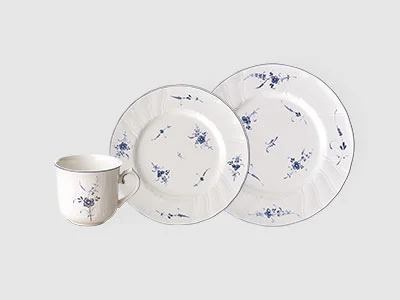 Image de la collection - Vieux Luxembourg Une tasse en céramique blanche et deux assiettes aux motifs floraux bleus sont disposées sur un fond sobre gris clair.