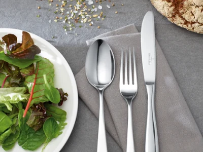 Couverts de table Sereno Une assiette de salade à côté d'une cuillère, d'une fourchette et d'un couteau sur une serviette grise sur une table grise.
