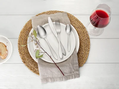 Des couverts de table Arthur sur une assiette blanche Artesano de Villeroy & Boch Une table dressée avec une assiette Artesano, des couverts de table Arthur et une serviette sur un set de table rond. À côté, sont posés un verre à vin rouge et une petite assiette avec des tranches de pain. Sur l’assiette se trouve une branche.