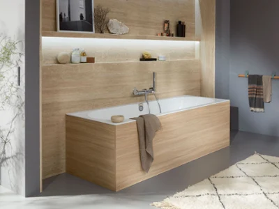 Une baignoire Oberon blanche avec un habillage en bois Une salle de bains moderne avec une baignoire carrée recouverte de bois, des étagères intégrées avec des décorations et des articles de toilette, une serviette suspendue sur le côté et une palette de couleurs douces et neutres.