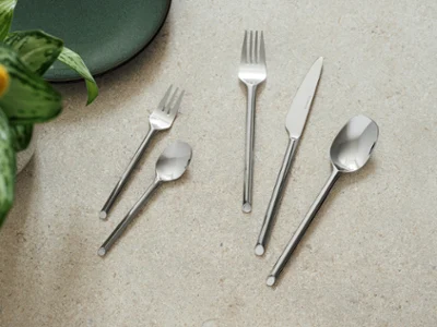Cinq couverts like de like.by Villeroy & Boch- fourchette, couteau, cuillère, fourchette à dessert et cuillère à café Cinq couverts like. Like. by Villeroy & Boch - fourchette, couteau, cuillère, fourchette à dessert et cuillère à café - sont disposés sur une surface en pierre claire à côté d'une assiette verte et d'une plante.