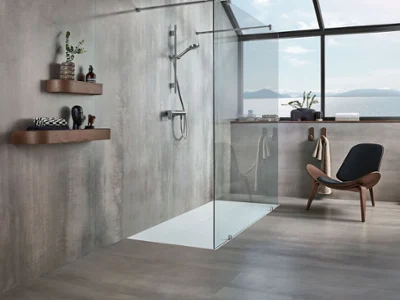 Un receveur de douche à l’italienne Squaro Infinity simple et blanc Une salle de bains moderne avec une douche vitrée et un grand receveur de douche plat blanc, des étagères en bois avec des articles de toilette, une chaise noire et une grande fenêtre avec vue sur la mer et les montagnes.