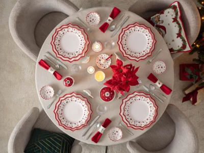 Table de Noël festive avec la vaisselle Toy's Delight Vue d'en haut d'une table ronde élégamment dressée pour Noël avec la vaisselle en porcelaine Toy's Delight de Villeroy & Boch, des serviettes rouges, des bougies et un centre de table avec un poinsettia rouge, à côté d'un sapin illuminé.