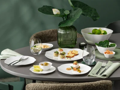 Une table ronde dressée avec la vaisselle blanche de la collection Pura. Une table ronde dressée avec la vaisselle blanche de la collection Pura, comprenant des assiettes à entrée, des verres, des serviettes et un centre de table composé de plantes vertes.