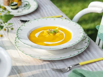 Assiette à soupe Colourful Spring avec soupe de patates douces et mangue Une assiette à soupe Colourful Spring avec une soupe de patates douces et de mangue, garnie d'herbes et de crème, est posée sur une table décorative en bois, à côté de laquelle se trouve une fourchette.