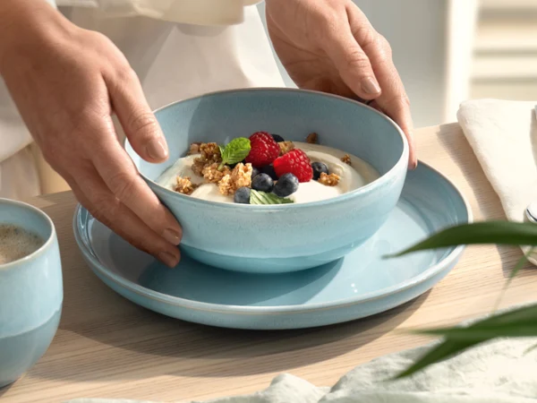 Des mains posant un bol bleu nacré Aqua rempli de yaourt sur une assiette bleue. Des mains déposant un bol bleu nacré Aqua rempli de yaourt, de muesli, de myrtilles et de framboises sur une assiette bleue.