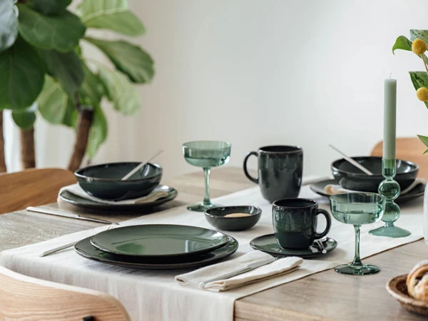 Une table à manger dressée avec Lave vert Une table à manger avec de la vaisselle verte, des verres, des couverts, une bougie et un vase avec des fleurs jaunes.