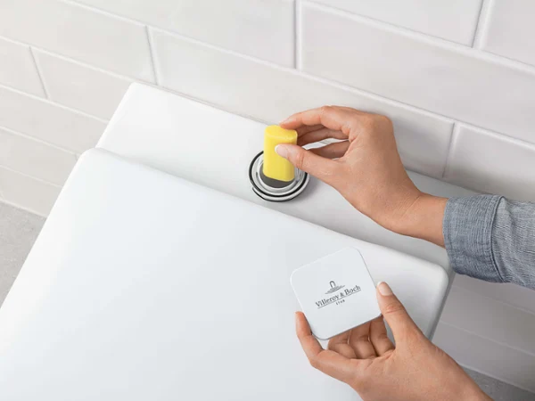 Remplissage du dispositif ViFresh de toilettes Une personne jette un bloc WC jaune dans le réservoir de chasse d’eau de toilettes. En arrière-plan, on peut voir des murs carrelés en blanc.