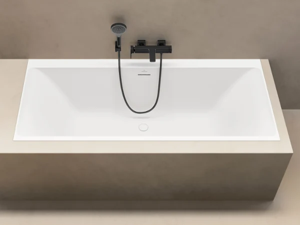 Une baignoire rectangulaire blanche minimaliste avec un robinet noir moderne et une douchette à main devant un mur beige.