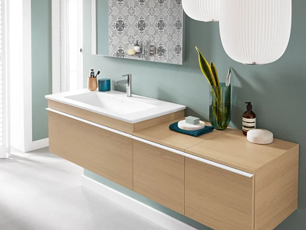 Une salle de bains moderne avec un plan de toilette suspendu en bois, un grand lavabo blanc Venticello, un mur de fond carrelé décoratif et des accessoires minimalistes, dont un vase vert avec une plante, du savon et des articles de toilette.