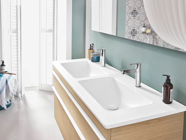 Une salle de bains moderne équipée d’un plan double avec des robinetteries argentées, un meuble en bois et des articles de toilette. La lumière naturelle pénètre dans la pièce par une fenêtre aux volets blancs.