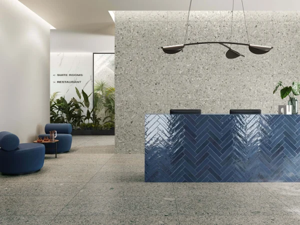 Code 2 Carreaux dans un lobby Lobby d'hôtel moderne avec une réception bleue structurée, un mobilier minimaliste, des lampes suspendues et un coin rempli de plantes vertes.
