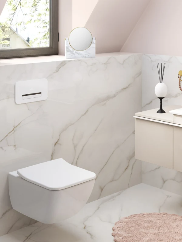 Une salle de bains moderne avec mur marbré, un lavabo suspendu avec un robinet doré, des toilettes suspendues, un miroir rond, un petit tapis et divers articles de toilette.