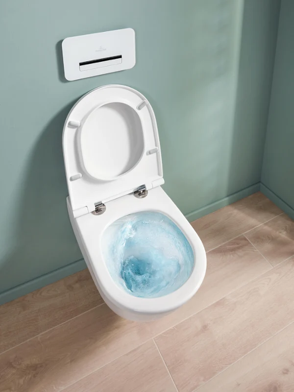 Universo TwistFlush, avec la chasse d’eau en fonctionnement, avec un mur vert clair Une cuvette de toilettes blanche ouverte avec de l’eau de chasse d’eau bleue, devant un mur bleu-vert et un sol en bois clair.
