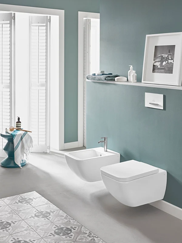 La salle de bains moderne aux murs bleu-vert dispose de WC suspendus Venticello et d’un bidet. On voit également une étagère avec des articles de toilette et une photo encadrée, ainsi qu’une petite table d’appoint avec une serviette et des ustensiles de bain à côté des volets.