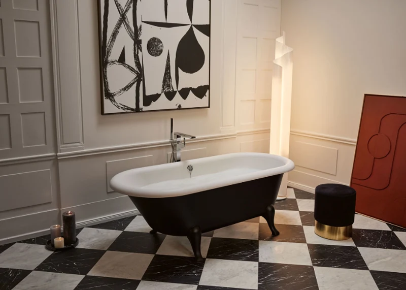 Une baignoire en îlot noire et blanche Hommage Une salle de bains élégante avec une baignoire en îlot Hommage noire et blanche, un carrelage de sol à carreaux, un tabouret foncé avec un pied doré, des bougies et une grande œuvre d’art géométrique.