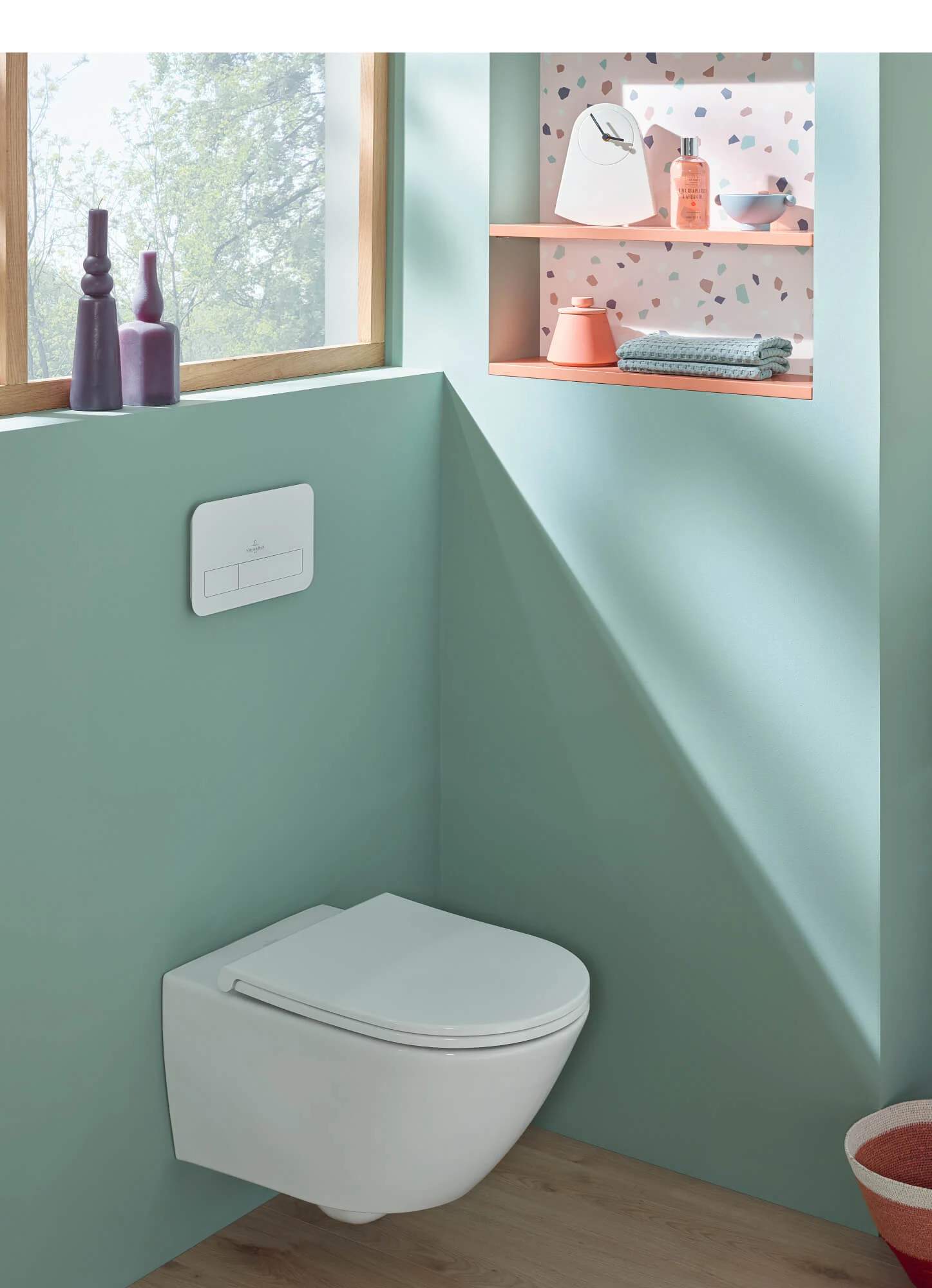 Une salle de bains moderne avec des toilettes suspendues et des étagères au-dessus, sur lesquelles sont posés des articles de toilette colorés. Une grande fenêtre laisse entrer la lumière naturelle.