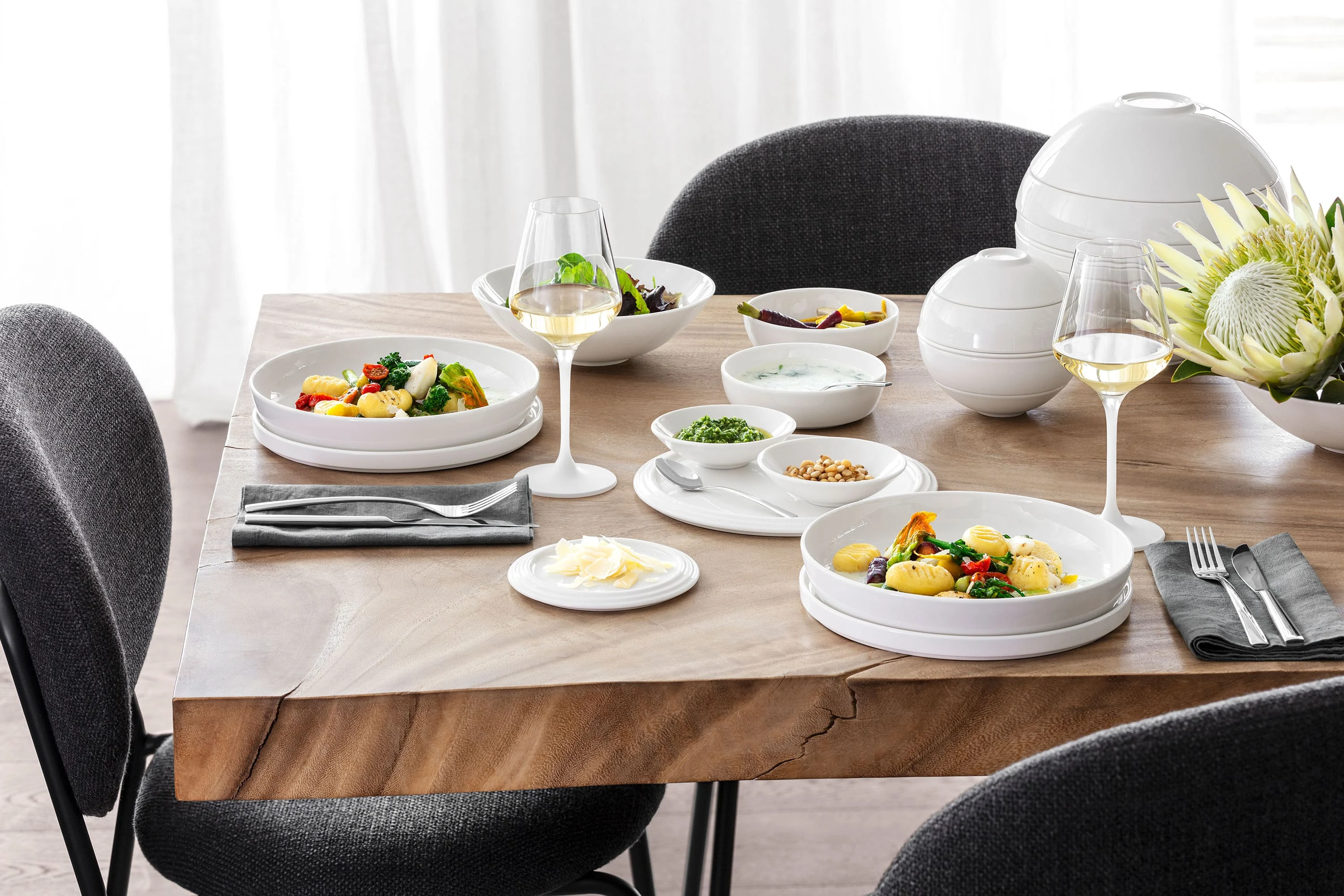 Une table dressée avec La Boule avec des chaises noires et grises tout autour Une table dressée La Boule avec deux assiettes pleines de nourriture, deux verres de vin blanc, différents bols avec des accompagnements et une composition florale avec des chaises noires et grises tout autour.