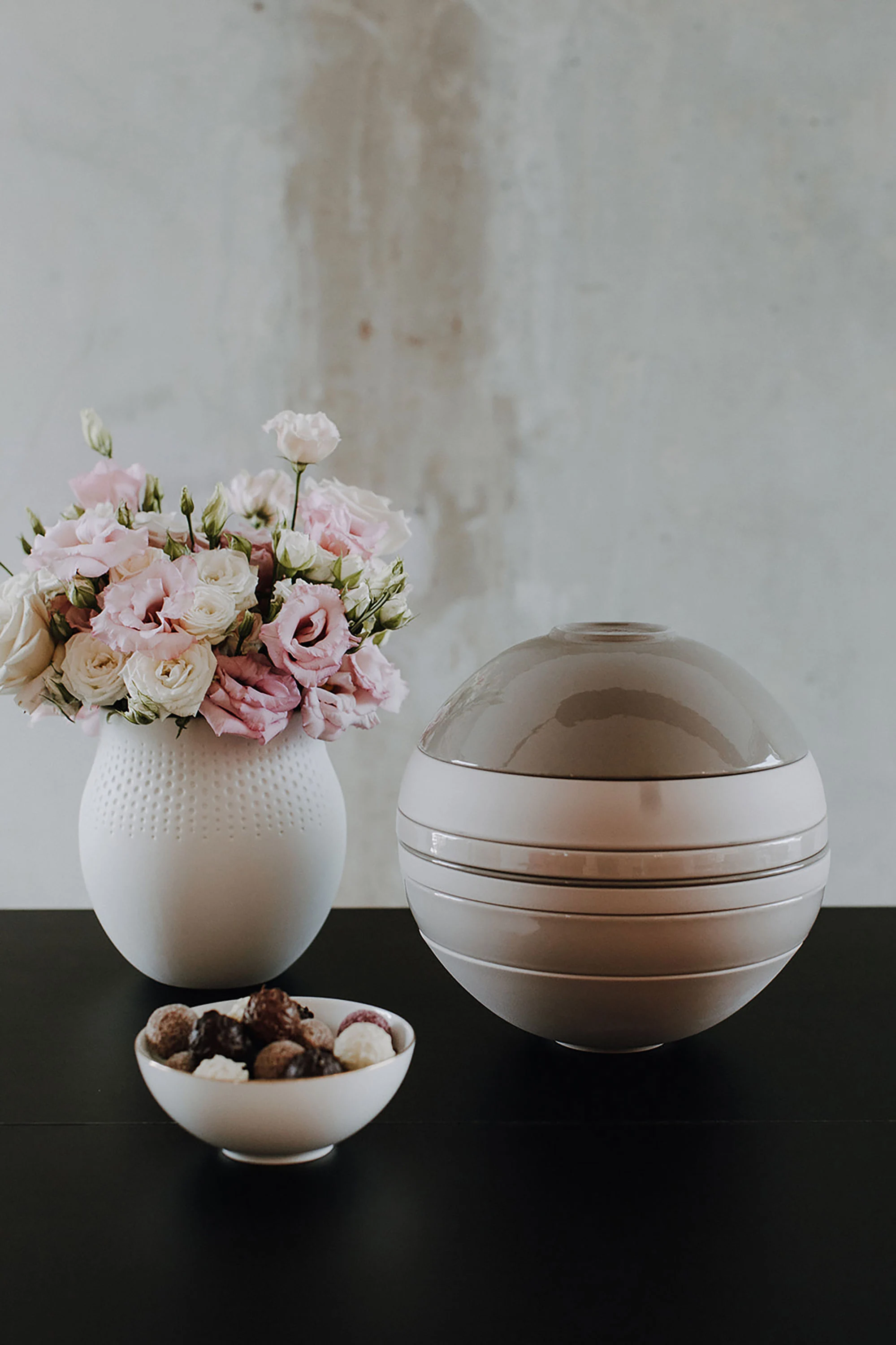 Un vase avec des fleurs roses, un ornement rond réfléchissant et une coupe avec différentes pierres, disposées sur une surface noire.