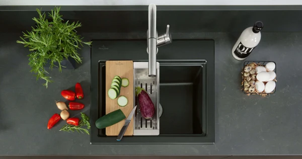 Un évier de cuisine Subway Style avec la robinetterie et les accessoires assortis sur un plan de travail sombre avec des légumes découpés et non découpés.