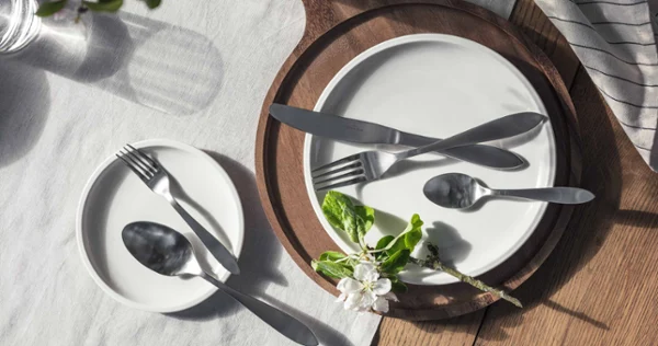 Une table dressée avec deux assiettes blanches, des couverts Arthur disposés dessus et une branche de fleur sur une nappe ensoleillée.