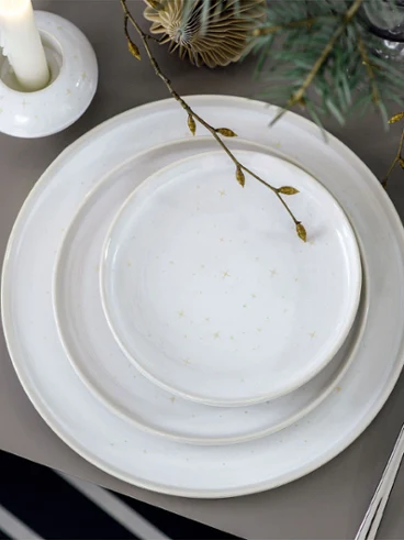Un dîner de fête avec Winter Glow à la lueur d’une bougie Une table festive, décorée avec des assiettes Winter Glow, des couverts de table et un bougeoir Winter Glow, entourée de branches de sapin.