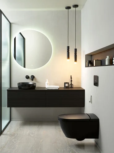 Une salle de bains moderne avec un miroir rond rétroéclairé, un plan de toilette noir suspendu avec une vasque à poser, des toilettes Antao noires de Villeroy & Boch et un décor minimaliste. Deux lampes suspendues sont accrochées au plafond. Une cloison en verre se trouve à gauche.