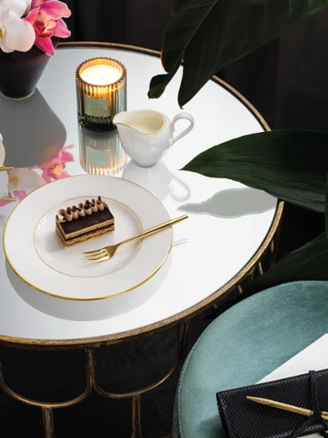 Une table ronde en miroir avec une part de gâteau sur une assiette gracieuse Une table ronde en miroir avec une part de gâteau sur une assiette de grâce, une tasse de café, une bougie, une plante et un tabouret bleu-vert à côté.