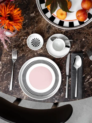 Une table ronde en marbre avec deux couverts Memphis, chacun avec une assiette, des couverts et une tasse. Un bol de pommes et un vase Memphis avec des fleurs ornent le centre.