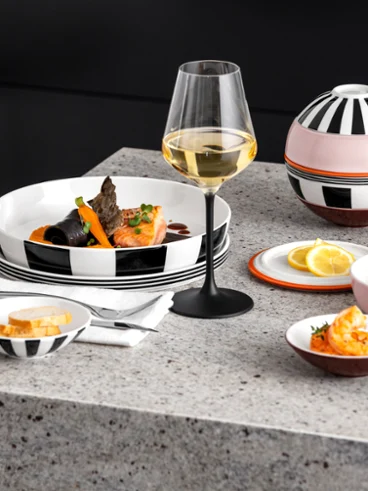 Fruits de mer garnis sur La Boule Memphis de Villeroy & Boch Une table dressée de façon moderne avec un verre de vin blanc, des fruits de mer servis, du pain tranché et des quartiers de citron sur un plan de travail en granit.