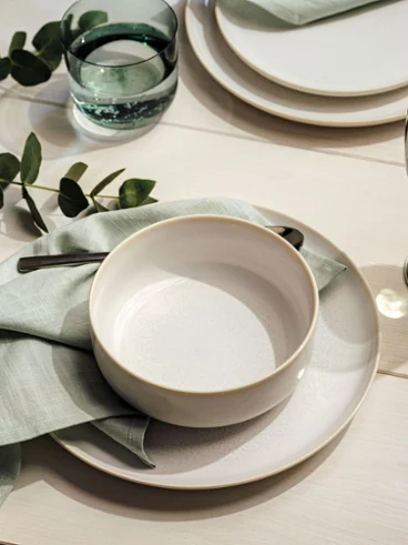 Un bol blanc Crafted sur une assiette Crafted Un Crafted Bol blanc sur une assiette Crafted recouverte de serviettes vert menthe et d'eucalyptus.