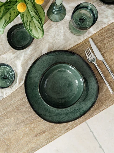 Table en bois élégamment dressée avec de la vaisselle en céramique "Lave Vert", des couverts en argent "like" de like. by Villeroy & Boch et des verres Like assortis sur une serviette en tissu crème. Une plante verte avec des accents jaunes complète l'arrangement raffiné.