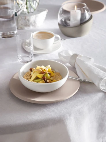 Un plat creux NewMoon contenant du porridge, rempli de fruits coupés et de noix, posé sur une table, accompagné d’un verre à eau et d’une tasse à café. Une bougie allumée et une serviette pliée sont également présentes.