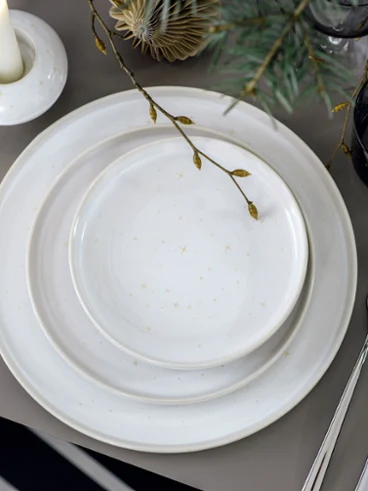 Un dîner de fête avec Winter Glow à la lueur d’une bougie Une table festive, décorée avec des assiettes Winter Glow, des couverts de table et un bougeoir Winter Glow, entourée de branches de sapin.