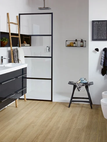 Une salle de bains de la collection Avento de Villeroy & Boch Une salle de bains moderne avec double lavabo, une douche avec un cadre noir, un sol en bois, des WC et un bidet suspendus, une horloge, une étagère en forme d’échelle et divers objets décoratifs.