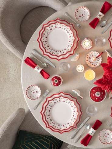Table de Noël festive avec la vaisselle Toy's Delight Vue d'en haut d'une table ronde élégamment dressée pour Noël avec la vaisselle en porcelaine Toy's Delight de Villeroy & Boch, des serviettes rouges, des bougies et un centre de table avec un poinsettia rouge, à côté d'un sapin illuminé.