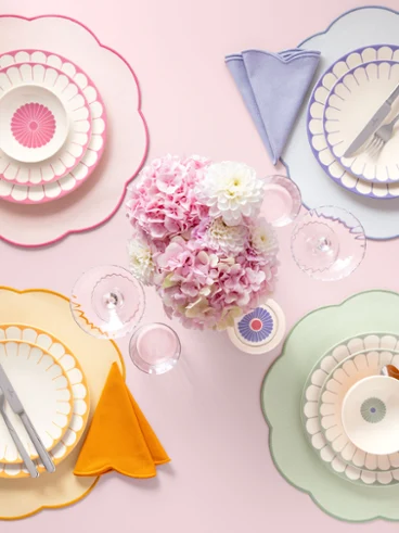 Quatre couverts aux couleurs pastel de la collection Fleur Quatre couverts aux couleurs pastel de la collection Fleur, avec des assiettes incurvées, des serviettes et des couverts, entourent un bouquet de fleurs roses.