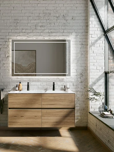 Une salle de bains moderne avec meuble suspendu en bois Finion, un grand miroir lumineux, des murs en briques et de grandes fenêtres. On y trouve un tabouret noir et de la décoration sur la tablette et le long du rebord de la fenêtre.