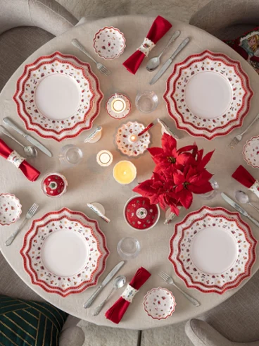 Table de Noël festive avec la vaisselle Toy's Delight Vue d'en haut d'une table ronde élégamment dressée pour Noël avec la vaisselle en porcelaine Toy's Delight de Villeroy & Boch, des serviettes rouges, des bougies et un centre de table avec un poinsettia rouge, à côté d'un sapin illuminé.