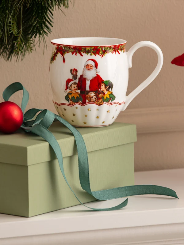 Annual Chistmas edition 2026 mug Une boîte cadeau verte avec un nœud, une décoration de Noël rouge et une tasse avec un motif de Père Noël de Villeroy & Boch devant un fond blanc, avec des décorations de Noël visibles en arrière-plan.