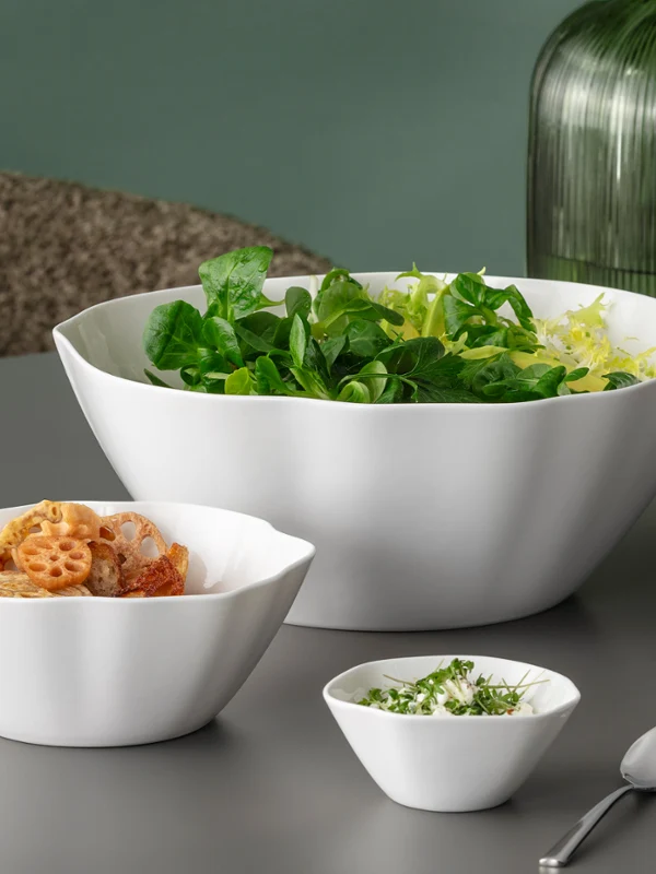 Trois bols blancs Pura avec salade, nouilles et herbes aromatiques Trois bols blancs Pura contenant de la salade, des pâtes et des herbes hachées sur une table grise, avec des vases verts en arrière-plan.