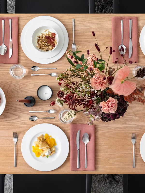 Mise en table florale avec Royal de Villeroy & Boch Une table rectangulaire en bois est dressée pour quatre personnes. Il y a des assiettes avec de la nourriture, des serviettes roses, des couverts, des bougies et une composition florale. Des raisins et des épices sont également présents.