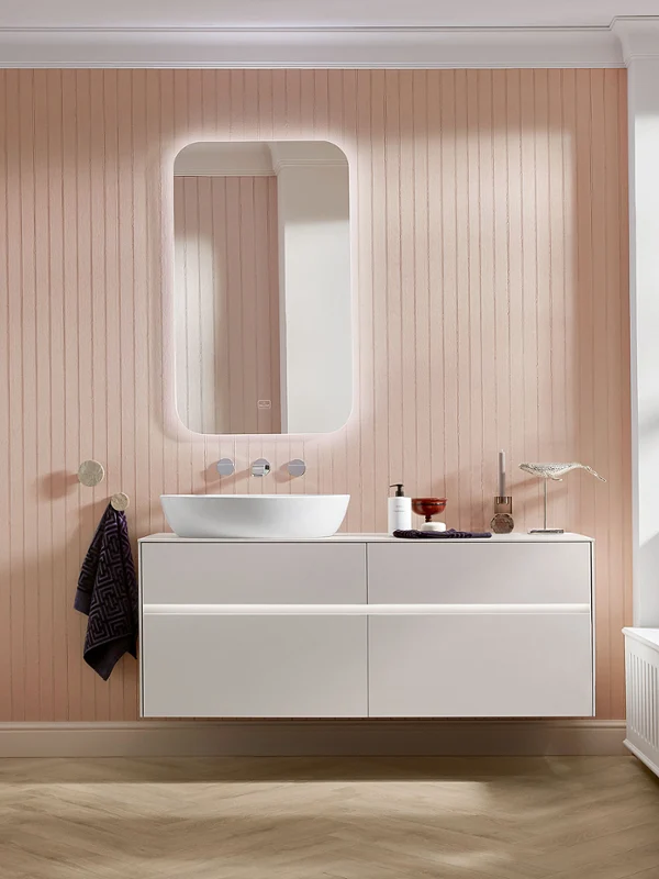 Lavabo Artis blanc sur meuble sous-vasque Collaro Salle de bains moderne avec un revêtement mural en bois, un meuble vasque suspendu, un lavabo rond, un miroir éclairé et des fleurs sur le rebord de la fenêtre.