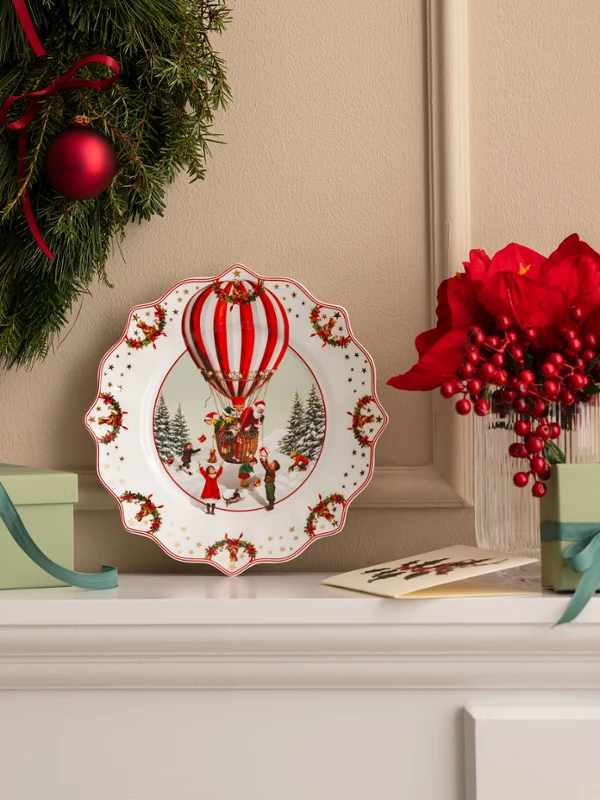 Annual Chistmas edition 2026 plate Une couronne de Noël, une assiette décorative ornée d'une montgolfière de Villeroy & Boch, des boîtes cadeaux vertes et des fleurs rouges, disposées sur une cheminée blanche.