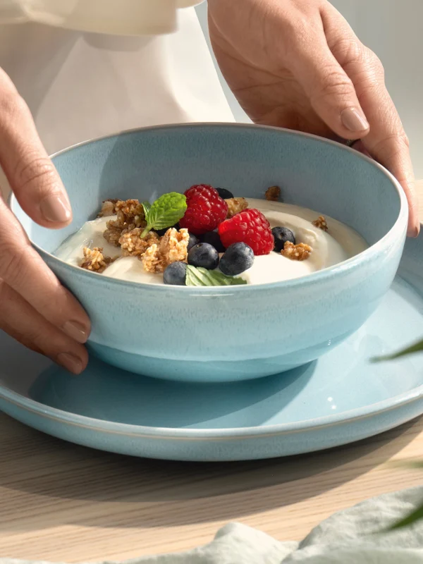 Des mains déposant un bol bleu nacré Aqua rempli de yaourt, de muesli, de myrtilles et de framboises sur une assiette bleue.