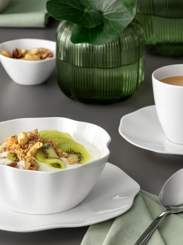 Une coupe Pura blanche avec des céréales et du kiwi et une tasse de café sur une soucoupe Un bol blanc Pura contenant du muesli et du kiwi, ainsi qu'une tasse de café posée sur une soucoupe, sur une table grise recouverte d'une serviette verte.