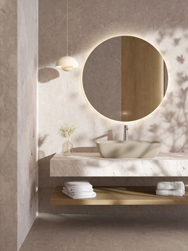 Lavabo Artis dans une salle de bain en marbre avec miroir rond Meuble de salle de bain minimaliste avec miroir rond rétroéclairé, vasque, serviettes et petit vase sur une étagère flottante.